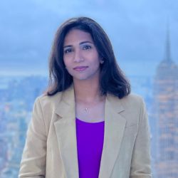 New Board of Directors member Tharsini Sivathasan, wearing a beige jacket and purple shirt. She is standing in front of a blurred cityscape.