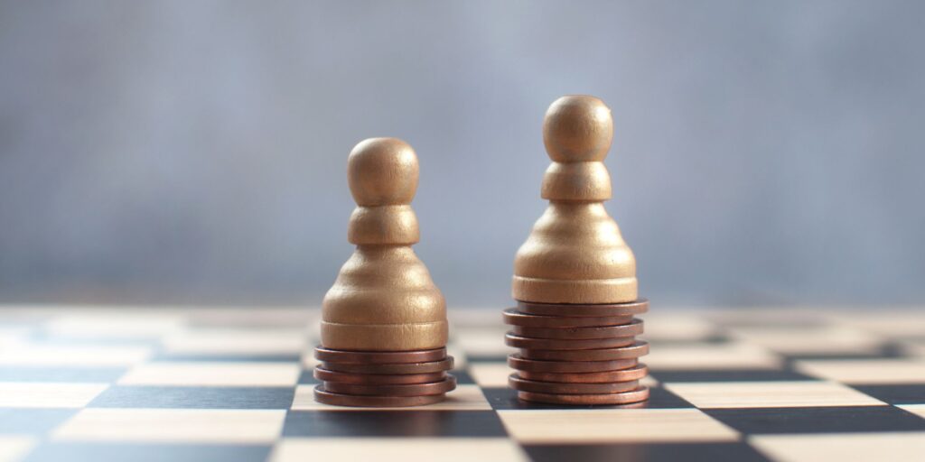 A close-up image of a chessboard with a focus on a golden pawn standing atop a stack of copper coins, while another pawn, also on a stack of coins, is in the background.