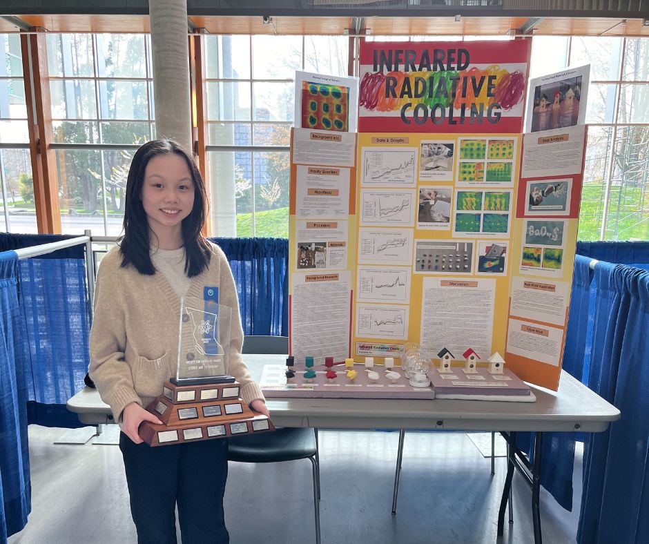 Stephanie standing in front of a science fair project titled 'Infrared Radiative Cooling', displaying a trophy and a table with various diagrams and models.